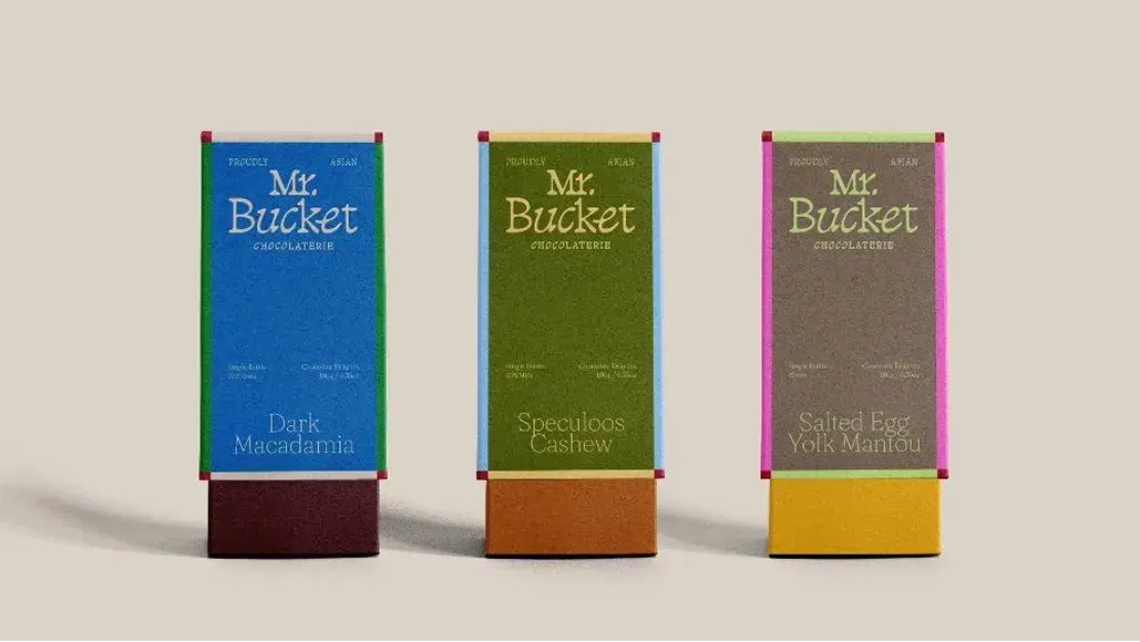 Three chocolate bars with colorful packaging labeled: Dark Macadamia, Speculoos Cashew, and Salted Egg Yolk Mantou by Mr. Bucket Chocolaterie.