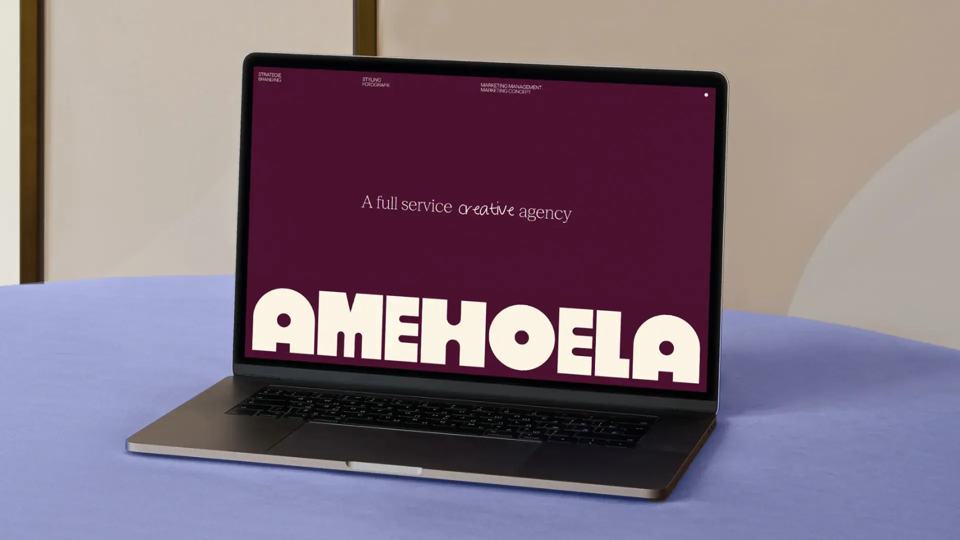 Laptop on a blue table displaying a website with maroon background and white text: "A full service creative agency AMEHOELA".