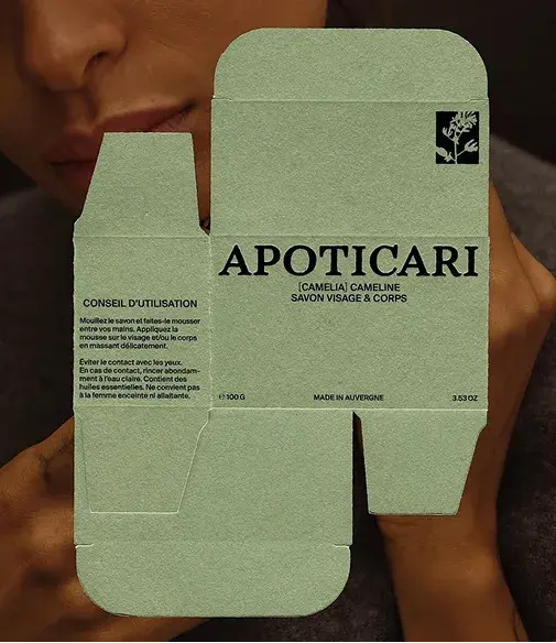 Person holding a green Apoticari soap packaging with text in French, featuring face and body usage instructions.