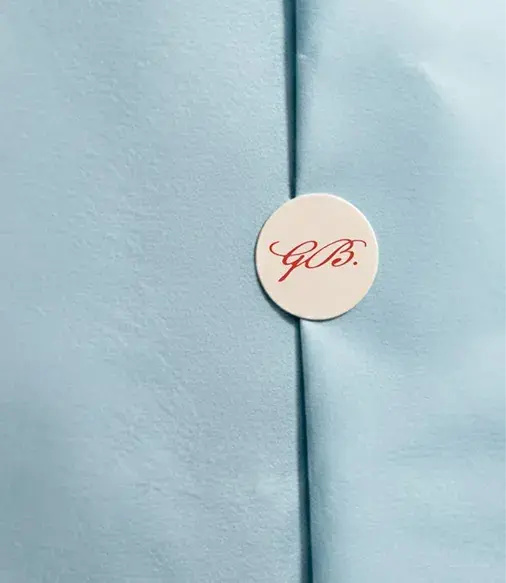 Close-up of light blue fabric with a small round sticker reading "G.B." in red script, placed over a seam.