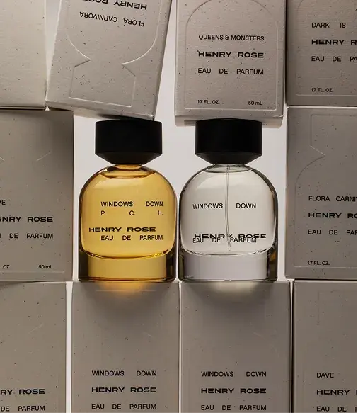 Two bottles of Henry Rose "Windows Down" perfume, one with amber liquid and the other clear, surrounded by stacked boxes.