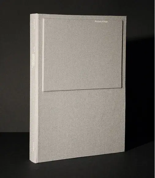 Gray book standing on a dark surface, featuring a minimalistic cover with a subtle raised rectangular design and small white text on the spine.