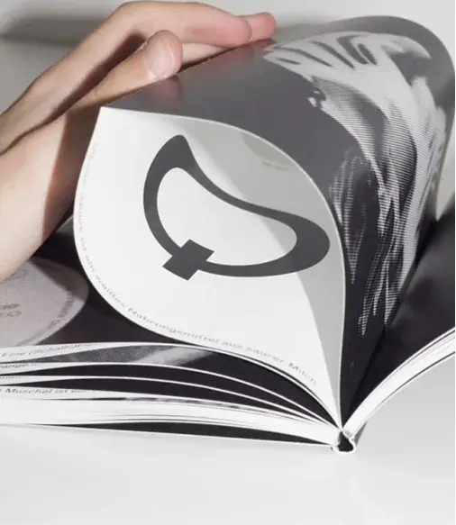 Hand flipping through a black-and-white magazine, displaying a page with a bold, abstract symbol.