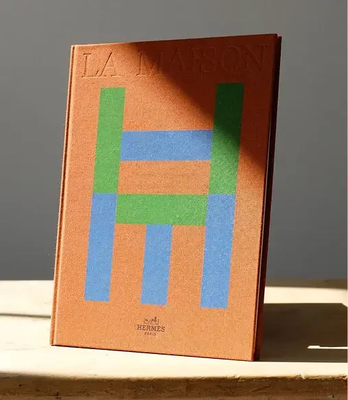 A book titled "LA MAISON" with an abstract H design on the cover, standing on a table in soft natural light.