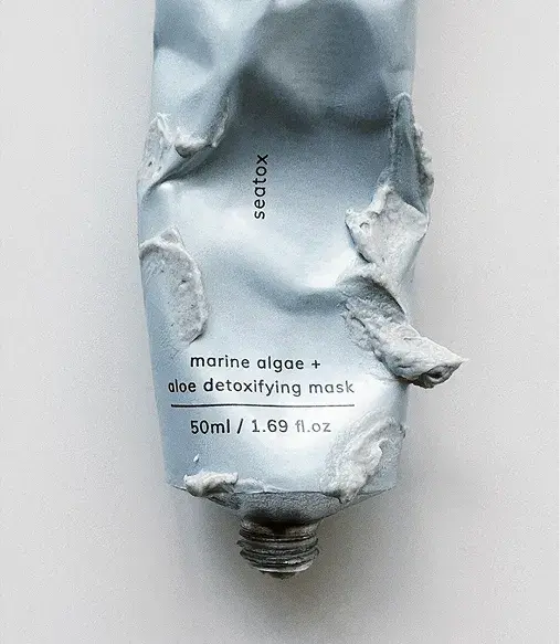 A crumpled light blue tube labeled "marine algae + aloe detoxifying mask," with some product oozing out. Capacity: 50ml (1.69 fl oz).