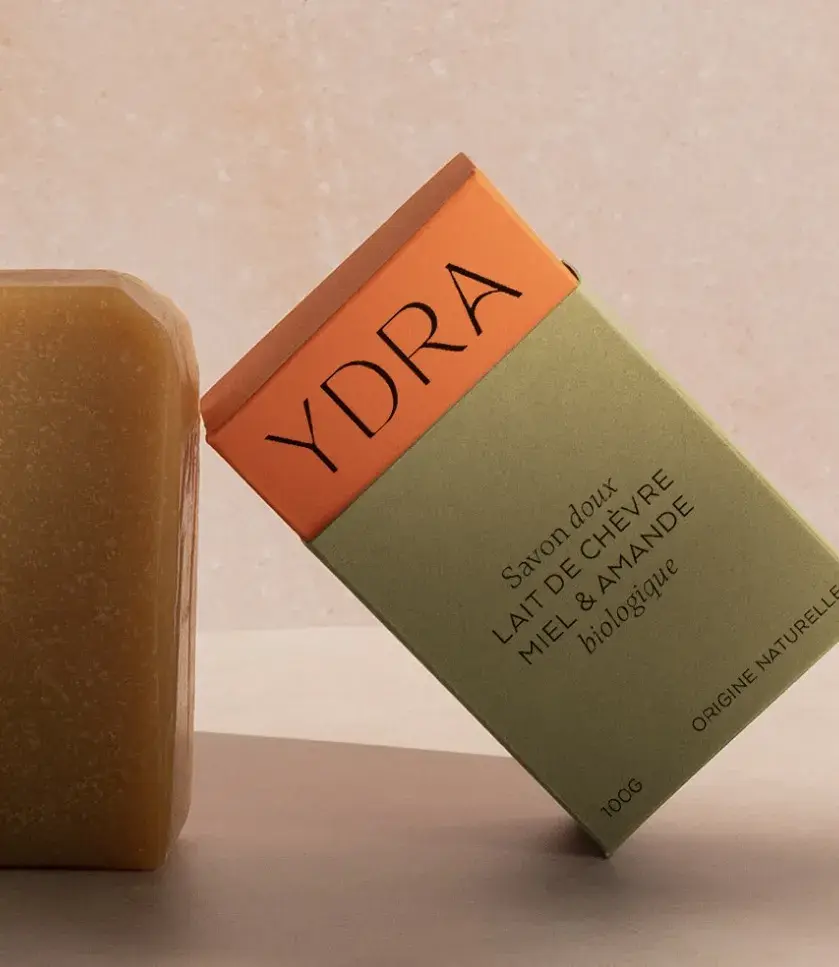A bar of soap next to a green and orange YDRA box labeled with "Lait de Chèvre Miel & Amande biologique."